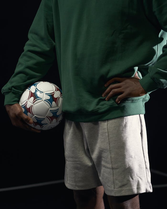 A person in sports attire holds a soccer ball in a dynamic pose on a dark background.